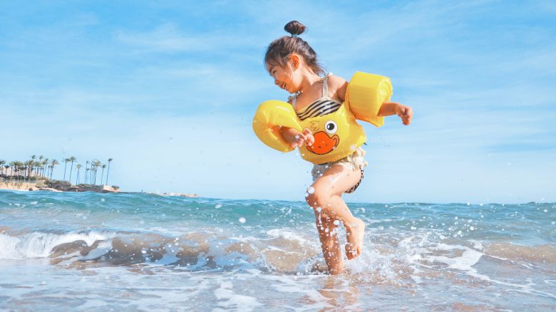 A young girl wearing inflatable arm floaties and a swimsuit is playing in the shallow waves of a beach under a blue sky with palm trees in the distance.