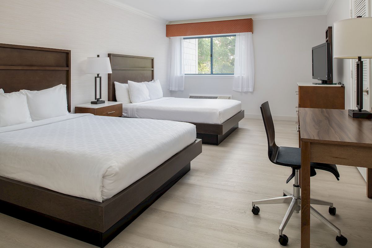 This image shows a hotel room with two double beds, a desk with a chair, and a TV. There's a window with white curtains.