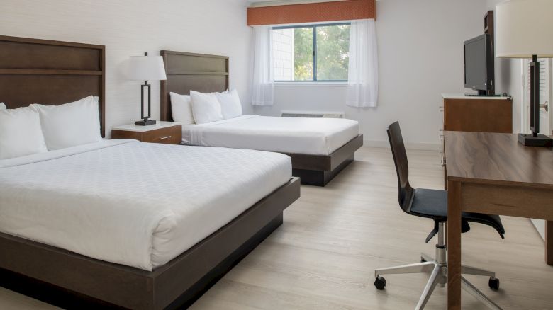 This image shows a hotel room with two double beds, a desk with a chair, and a TV. There's a window with white curtains.