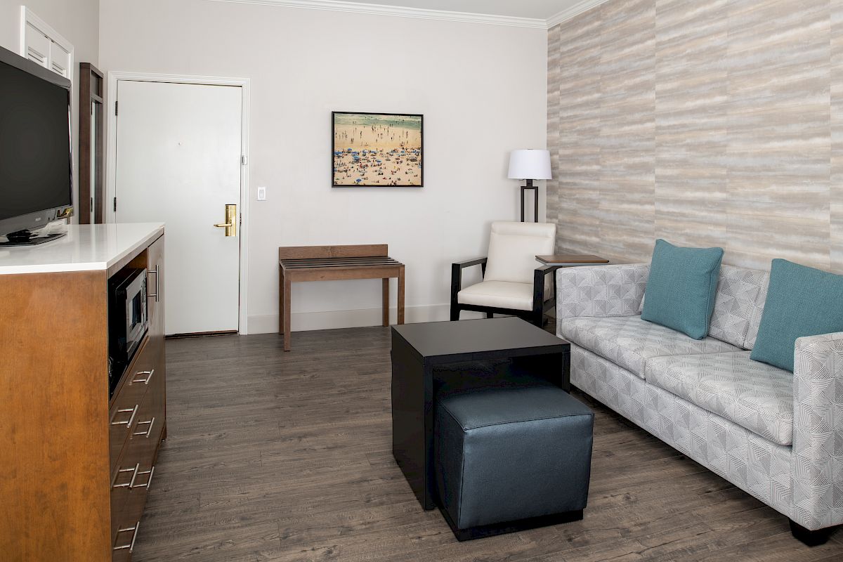 A modern hotel room with a sofa, TV, table, chair, and wall art. The room has neutral decor and wood flooring.