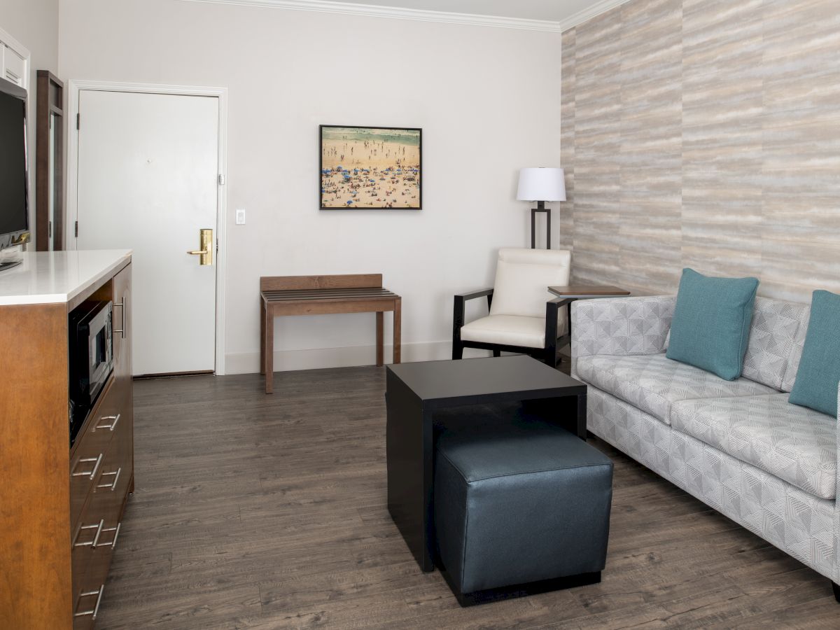 A modern hotel room with a sofa, TV, table, chair, and wall art. The room has neutral decor and wood flooring.