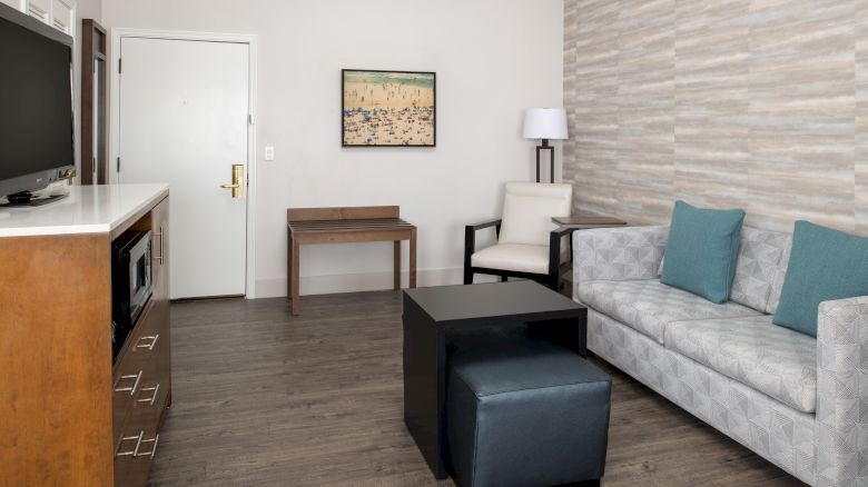 A modern hotel room with a sofa, TV, table, chair, and wall art. The room has neutral decor and wood flooring.