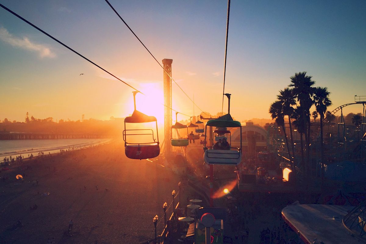 The image shows people on a cable ride at sunset near a beach, with palm trees and a pier in the background, creating a serene and picturesque scene.