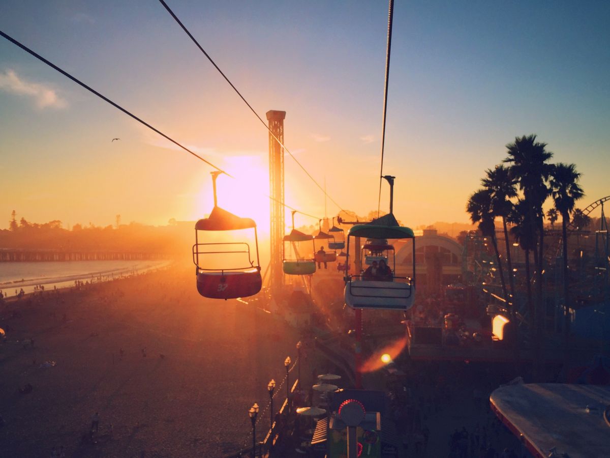 The image shows people on a cable ride at sunset near a beach, with palm trees and a pier in the background, creating a serene and picturesque scene.