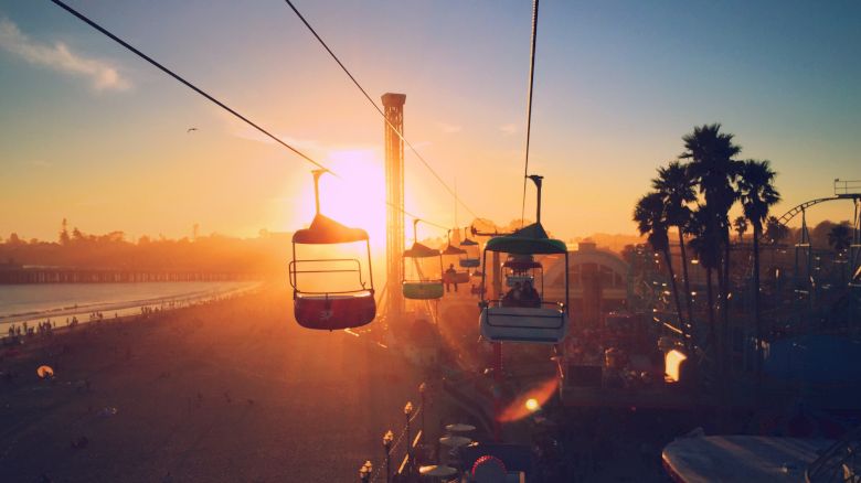 The image shows people on a cable ride at sunset near a beach, with palm trees and a pier in the background, creating a serene and picturesque scene.