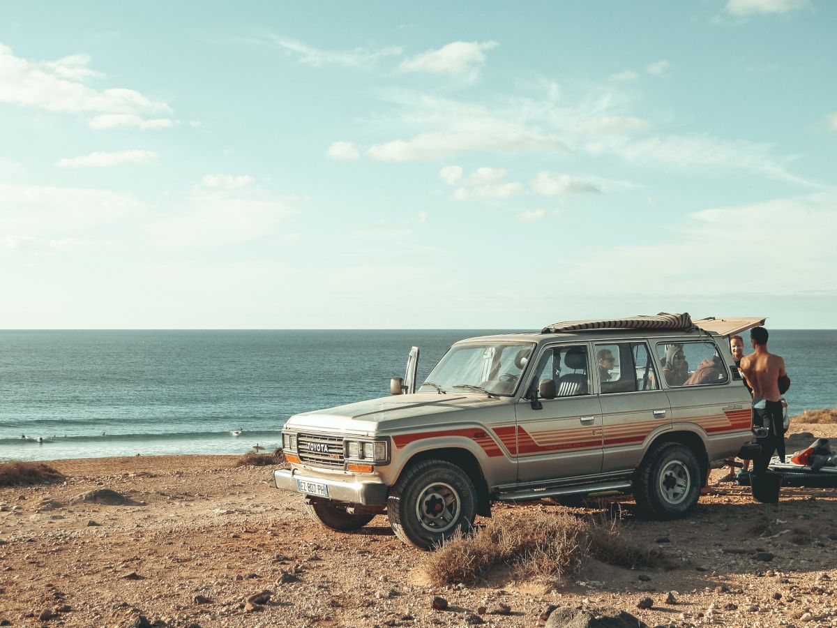 A beach scene with a SUV parked on sandy terrain, a shirtless man near the vehicle, and another car in the background. The ocean is visible.