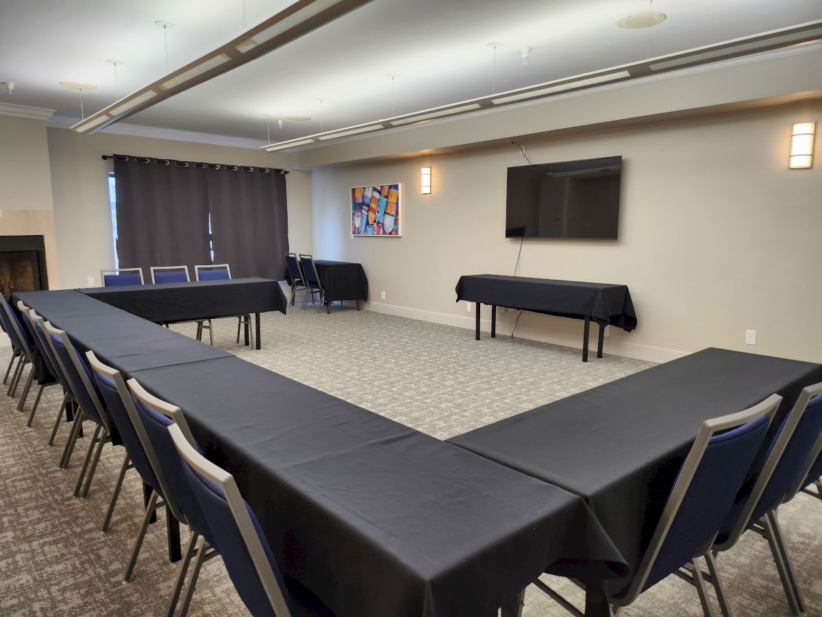 A conference room with U-shaped table setup, black tablecloths, blue chairs, a wall-mounted TV, a painting, and a closed curtain.