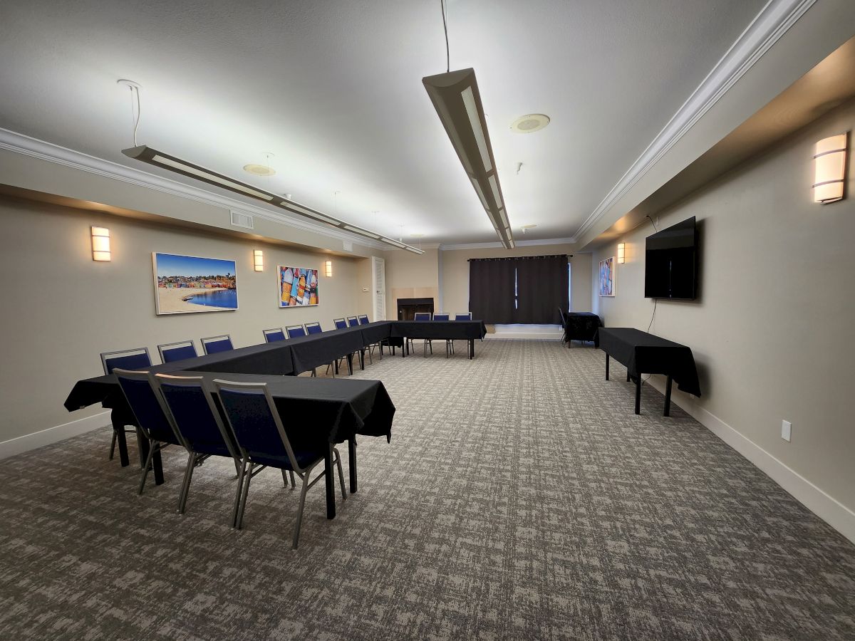 A conference room with a U-shaped table setup adorned with black tablecloths, chairs, wall paintings, a TV, and carpeted flooring.