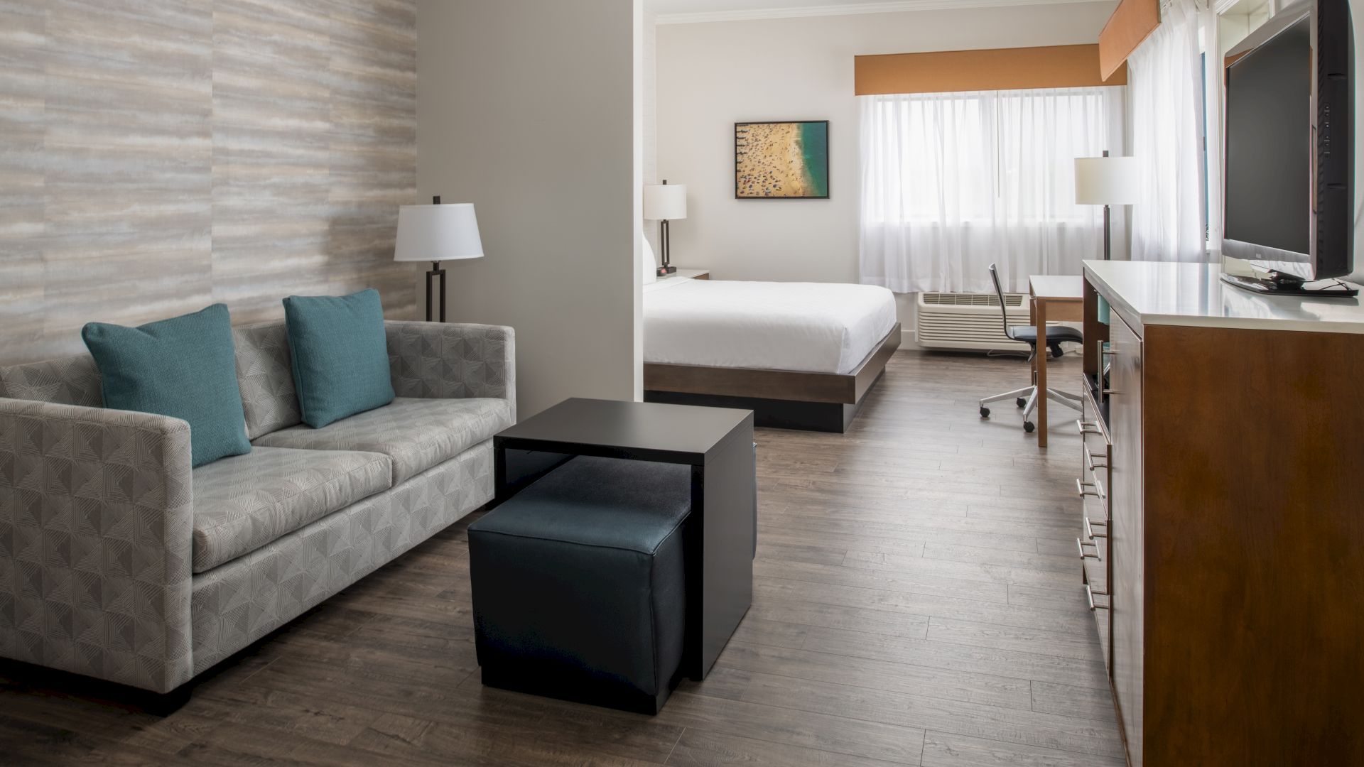 The image shows a modern hotel room with a sofa, coffee table, bed, TV, lamps, desk, chair, and artwork on the wall, all in neutral tones.