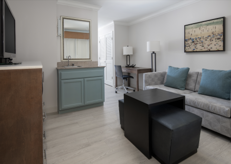 A modern living room with a sofa, TV, desk, and kitchenette, decorated with a wall painting and light blue accents.