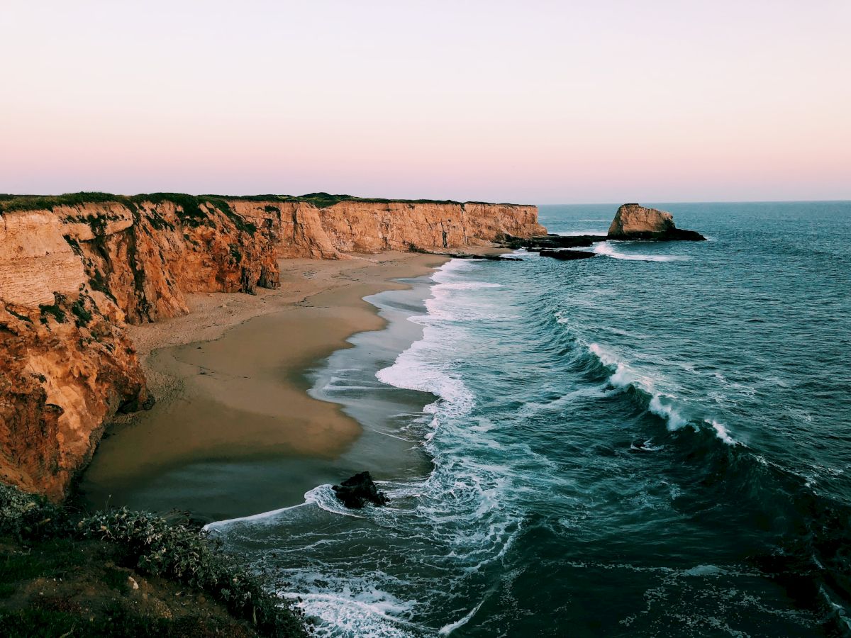 A scenic coastal view with cliffs, sandy beach, and gentle waves under a clear pinkish sky.