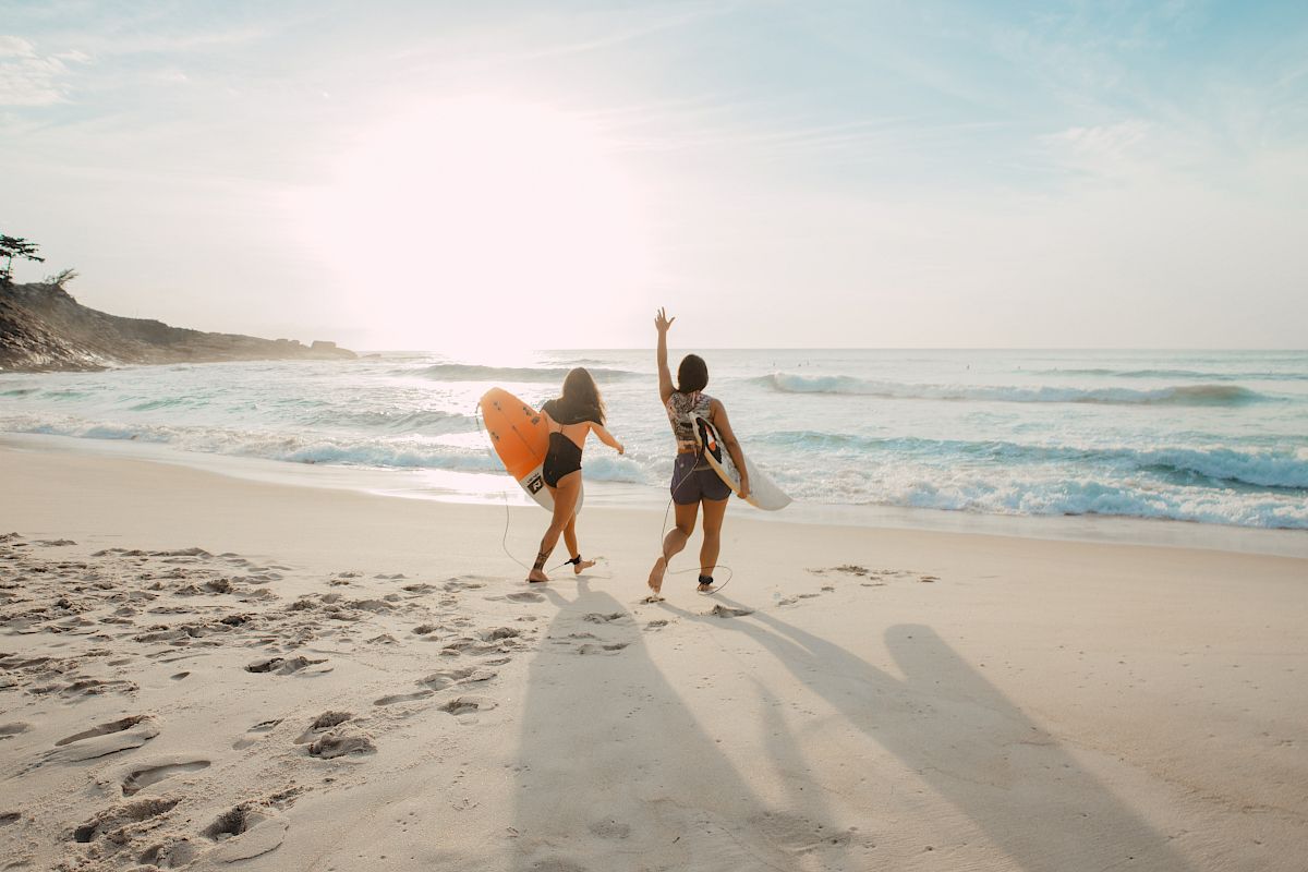 Two people with surfboards walk on a beach towards the ocean, one waves at the setting sun creating a warm, serene scene.
