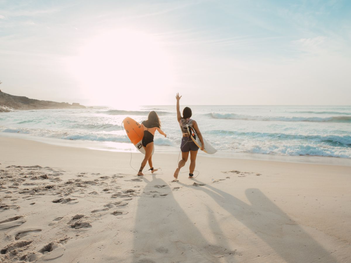 Two people with surfboards walk on a beach towards the ocean, one waves at the setting sun creating a warm, serene scene.