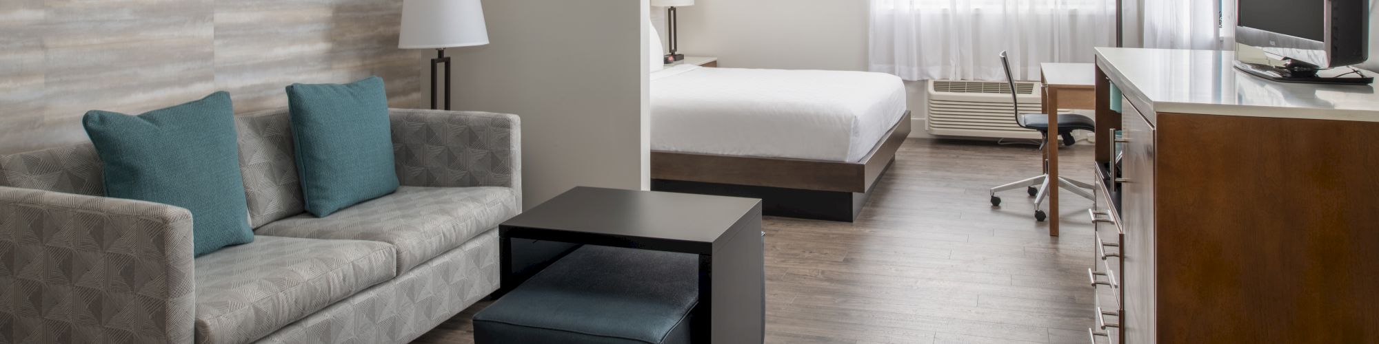 The image shows a modern hotel room with a sofa, coffee table, bed, TV, lamps, desk, chair, and artwork on the wall, all in neutral tones.