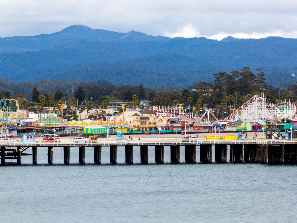 A lively seaside pier with colorful carnival rides, stalls, and tents along the water, backed by hills and a cloudy sky.