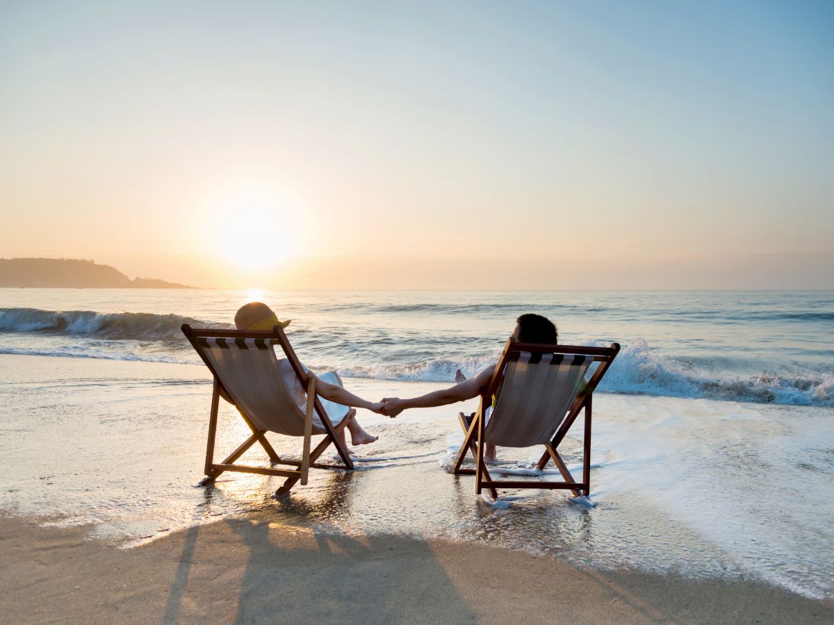 Two people sit in lounge chairs on the beach at sunset, holding hands as gentle waves wash ashore.